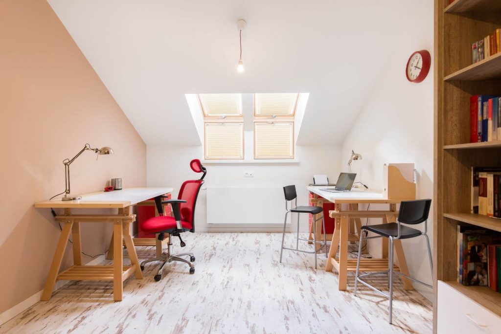 Home office with two wooden desks, skylight windows, and minimalist decor. The room features a mix of red and black chairs, whitewashed flooring, and built-in bookshelves for storage.