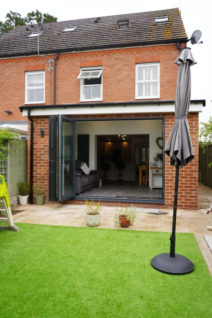 Rear extension on a red-brick home with dark-framed bifold doors opening onto a garden with artificial grass. The modern open-plan interior includes a living area and kitchen, seamlessly connected to the outdoor space.
