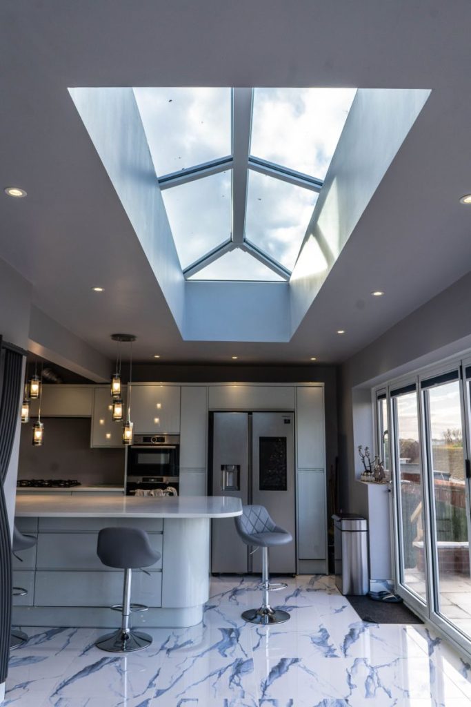 Modern kitchen extension featuring a large central roof lantern that floods the space with natural light. The sleek interior includes a glossy island unit, marble-effect flooring, and full-height sliding doors to the outside.